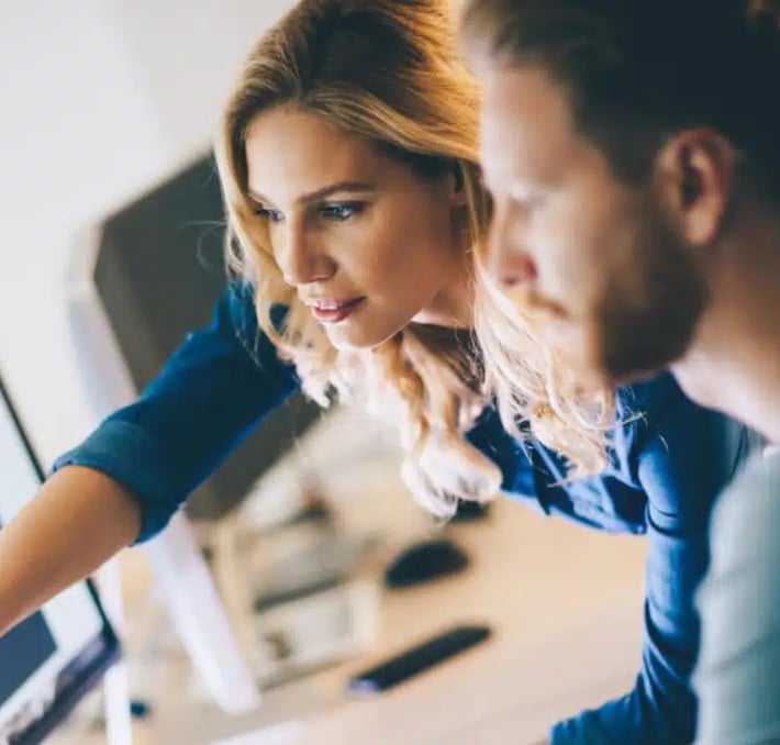 Two people in an office setting look at a computer monitor, with one pointing at the screen as they discuss CPQE and PLM solutions.