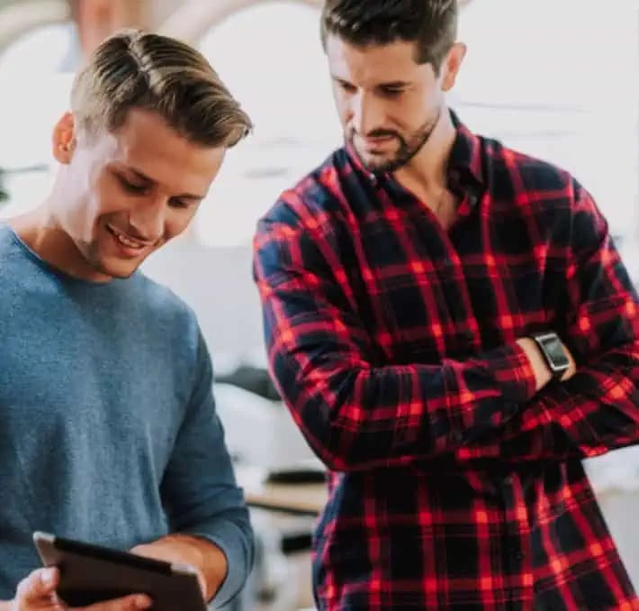 Two men stand indoors looking at a tablet together; one man is smiling and holding the tablet, while the other stands beside him with arms crossed, observing.