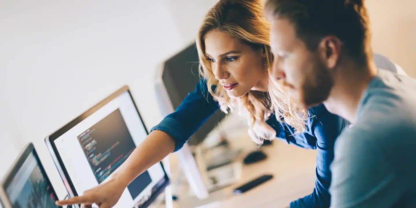 Two people work at a desk, looking at computer screens with code visible, as one person points at the monitor.