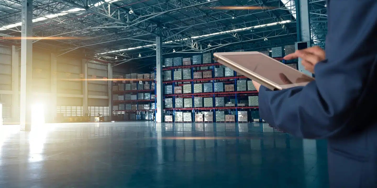 Person using a tablet in a large warehouse with high shelves full of boxes and pallets, illuminated by sunlight through open doors.