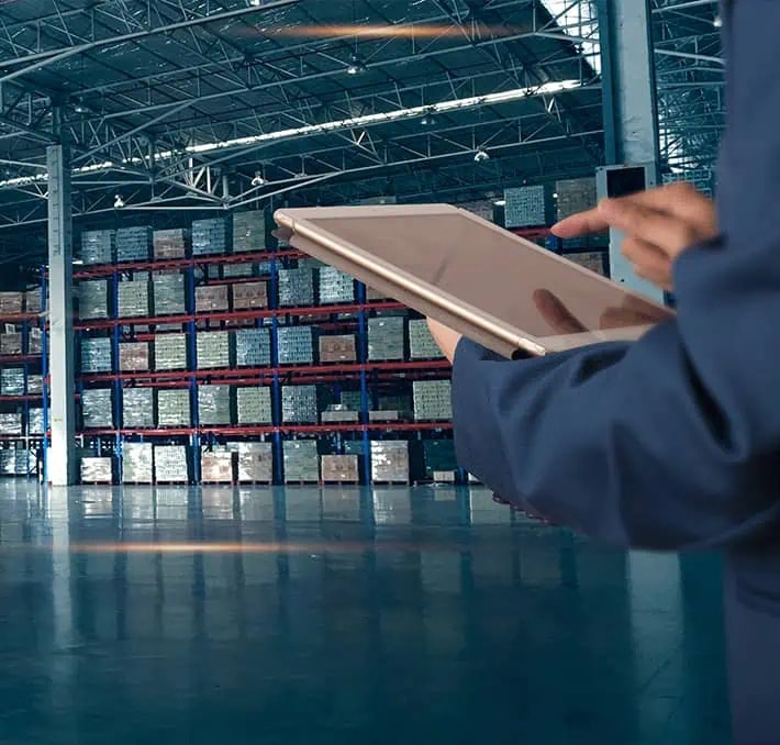 Person using a tablet in a large warehouse with stacked shelves filled with boxes and goods in the background.