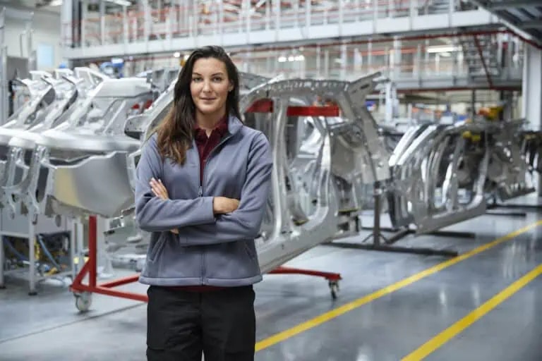 A woman stands with arms crossed in a modern automotive manufacturing facility, with metal car frames visible in the background.