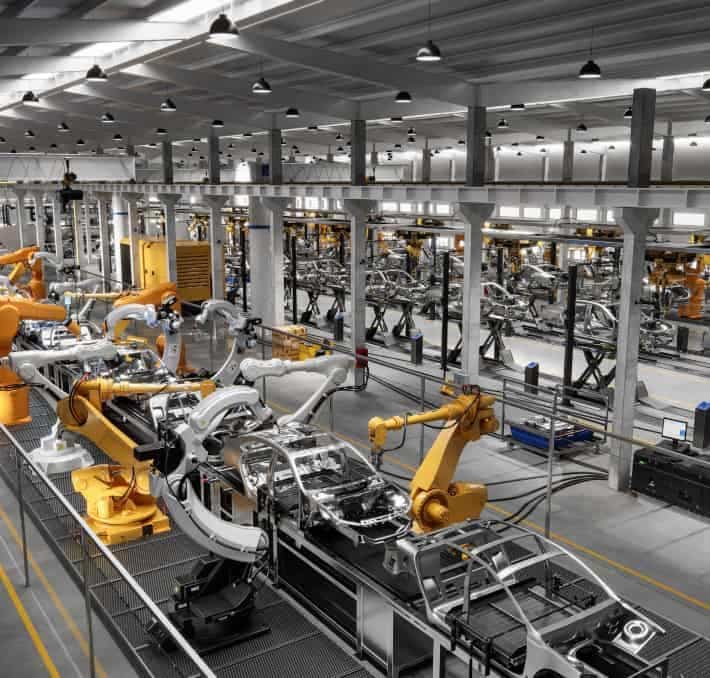 Robotic arms assemble car bodies on production lines in a spacious, well-lit automotive manufacturing factory.