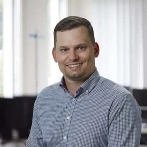 Man wearing a checked shirt is smiling at the camera in an office setting with large windows and white blinds in the background.
